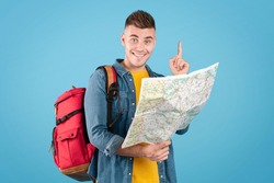 Positive millennial hiker with rucksack and map having idea about his camping trip on blue studio background. Portrait of cheerful young backpacker planning creative travel route