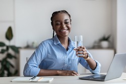 Hydration At Work. Happy Black Female Worker Holding Glass Of Mineral Water While Sitting At Workplace In White Office, Feeling Healthy And Motivated, Smiling At Camera, Copy Space