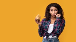 Cheat Meal. Portrait Of Happy African American Casual Woman Eating Tasty Burger, Licking Finger, Looking Away At Copy Space. Satisfied Woman Holding Fastfood, Isolated Over Orange Background, Banner