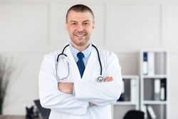 Professional Help. Portrait of smiling confident mature general practitioner standing with folded arms at his office in clinic and posing, looking at camera. Doctor wearing stethoscope and white coat