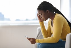 Upset african woman looking at cellphone screen while sitting on sofa at home, received bad news, sad black female reading unpleasant message or waiting for important call, side view with copy space