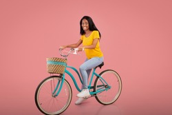 Full length portrait of beautiful African American woman riding vintage bicycle over pink studio background. Positive black lady on bike going cycling, enjoying her ride. Active lifestyle concept