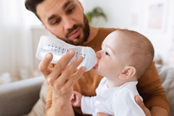 Fatherhood. Young Father Giving His Little Baby Toddler Bottle Of Water Feeding Him Sitting On Sofa At Home. Paternity Leave, Child Care, Parenting And Parenthood Concept. Selective Focus