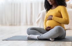 Pregnant african american woman sitting on yoga mat in living room, hugging her belly, copy space. Unrecognizable black expecting lady doing sport at home. Healthy lifestyle during pregnancy