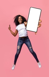Happy african american young woman showing empty smartphone screen while jumping up over pink studio background, collage, full size photo. Excited black lady recommending nice mobile application