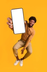 Excited black guy holding smartphone, showing blank screen, jumping up over orange studio background. Handsome african american young man recommending new mobile application, collage