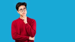 Good Offer. Portrait of pensive asian man in glasses looking up at copy space, touching his chin, isolated over blue studio background. Thoughtful teen guy thinking and making decision