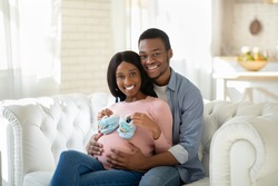 Black pregnant woman with tiny baby boties and her happy husband hugging on sofa at home. Future mom and dad with little shoes expecting baby, embracing on couch indoors