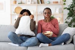 Frightened black girlfriends watching horror together at home, eating popcorn. Scared african american women sitting on sofa, hiding behind pillow and covering mouth with palm, watching movie