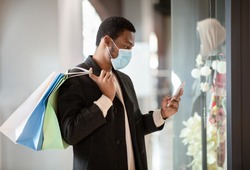 New normal, shopping and sales in Black Friday during COVID-19 pandemic. Serious young african american man in coat and protective mask holds colored bags and looks in smartphone near shop window