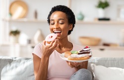 Young black woman on diet having cheat meal day, holding plate of sweets, stuffing her mouth with donut at home. African American lady eating unhealthy dessert. Poor nutrition concept