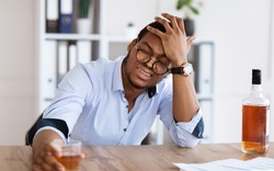 Things getting worse, difficulties at job. Frustrated young african american businessman drinking cause of having stress, holding glass of whiskey, holding his head, office interior, copy space