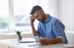 Worried male freelancer looking at smartphone screen while working at home office, received bad news, got scam message, thoughtful man touching forehead with concerned face expression, copy space