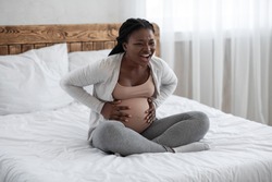 Pregnant black lady having labor contractions at home, suffering from acute belly ache and painful abdominal cramps, sitting on bed and touching her pregnant tummy, feeling unwell, free space