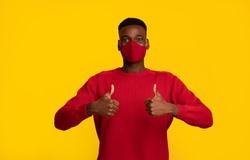 Happy black guy in red protective face mask showing thumbs up at camera, posing isolated on yellow background, african american man support social-distancing during coronavirus pandemic, copy space