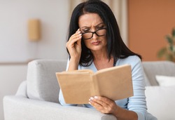 Portrait of mature woman touching glasses trying to read book, having difficulties seeing text because of vision problems. Negative emotion facial expression feelings reaction, health issues