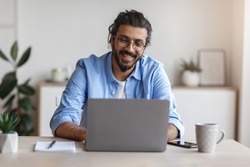 Freelance Work. Happy Millennial Indian Man Working On Computer At Home Office, Sitting At Desk With Laptop, Handsome Western Guy Looking At Device Screen And Smiling, Enjoying Remote Job, Copy Space