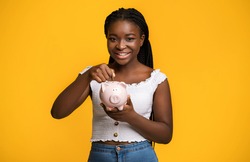 Economy Concept. Smiling Black Woman Holding And Inserting Coin At Piggy Bank, Saving Money For Future, Making Smart Investment, Positive Lady Standing Isolated Over Yellow Background, Copy Space
