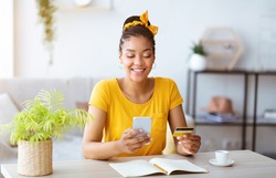 Online Order And Ecommerce Concept. Smiling African American Woman Using Smartphone And Holding Credit Card, Sitting At Desk With Open Notebook In Living Room At Home. Internet Shop Offers And Sales