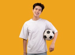 Football Player. Portrait of confident asian guy posing with soccer ball, standing isolated over pastel orange studio background, banner. Smiling young man in white t-shirt looking at camera