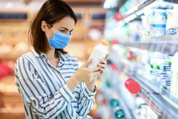 Choosing Milk. Female customer in medical surgical mask holding bottle of milk or yoghurt from the fridge, looking at dairy products, standing near refrigerator aisle, checking expiry date