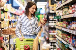 Portrait Of Smiling Woman With Shopping Cart In Supermarket Buying Groceries Food Walking Along The Aisle And Shelves In Grocery Store, Holding Glass Jar Of Sauce, Choosing Healthy Products In Mall