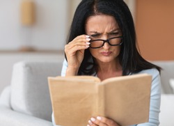 Portrait of confused senior woman squinting to see more clearly, wearing and touching eyeglasses, trying to read paper book, having difficulties seeing text because of bad vision problems and issues