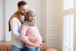 Happy Parents-To-Be. Lovely Muslim Couple Waiting For A Baby, Embracing Standing In Living Room At Home. Arabic Husband Hugging Pregnant Wife. Empty Space For Text