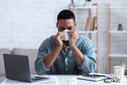 Sick Black Guy Working In Office, Sitting At Laptop And Blowing Nose In Paper Napkin Indoors. Ill Employee Concept. African Man Having Cold, Rhinitis And Sinusitis Symptoms At Work.