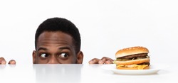 Funny Hungry African Guy Looking At Tasty Burger On Desk Having Food Craving Posing In Studio On White Background. Overeating Habit, Dieting And Nutrition, Cheat Meal Concept. Panorama