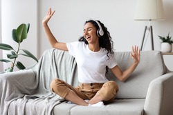Carefree brunette woman listening to music and singing, using headphones, sitting on couch in living room, copy space. Happy young lady with closed eyes enjoying music, home interior
