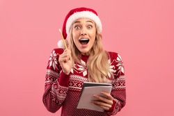 Excited young blonde woman in Christmas sweater and Santa hat writing New Year resolutions in notepad over pink studio background. Positive millennial lady making Xmas wish list