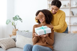 Happy celebration and congratulations from friend, idea for present and birthday. Smiling cute millennial mixed race lady closes eyes and gives gift to african american woman in living room interior