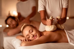 Couple Enjoying Herbal Massage. Relaxed Husband And Wife At Exotic Spa Resort, Lying With Eyes Closed Receiving Thai Massage With Aromatic Bags. Body Beauty Treatment, Relaxation And Wellness