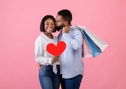 Passionate black man holding shopping bags, kissing his girlfriend with red heart on pink studio background. Loving guy and his girlfriend purchasing presents for Valentine's Day