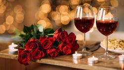 Romantic Dinner. Bouquet of flowers lying on the table, selective focus on bunch of roses, two glasses of red wine and candles on the wooden desk. Date concept, blurred background, banner, copy space