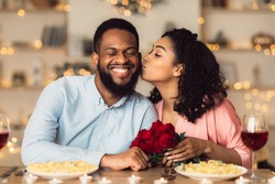 Celebrating Special Day. Beautiful black girlfriend kissing her smiling boyfriend in cheek, holding bouquet of red roses. Couple sitting at table in luxury restaurant during romantic date and dinner