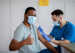 African American man in antiviral mask gesturing thumb up during coronavirus vaccination, approving of covid-19 immunization. Young doctor giving vaccine injection to male patient