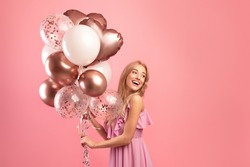 Party time. Charming blonde woman in stylish dress holding bunch of birthday balloons over pink studio background, copy space. Joyful young lady having fun celebration, enjoying holiday
