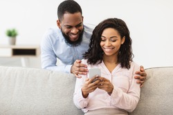 Cheerful millennial black couple browsing internet on smartphone, watching video together on gadget. Handsome boyfriend embracing his girlfriend from back, lady sitting on the couch