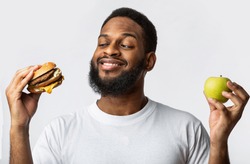 Black Guy Holding Burger And Apple Choosing Between Healthy Vs Unhealthy Food Standing In Studio On White Background. Male Nutrition And Junk Food, Cheat Meal On A Diet Concept