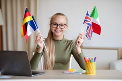 Cheerful blonde teen girl with glasses sitting at workdesk with international flags, home interior, using laptop, studying foreign languages. International education, online language courses