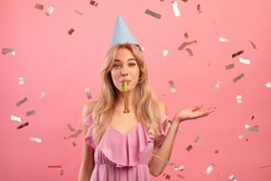 Gorgeous young woman in birthday cap blowing party horn on pink studio background with falling confetti. Positive millennial lady celebrating holiday. Festive concept