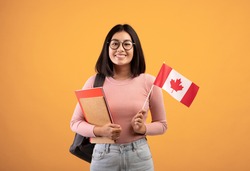Travel, modern education and student exchange. Young cheerful asian woman in glasses with notebooks, backpack holding small flag of Canada, isolated on beige background, studio shot, empty space
