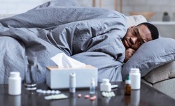 Sick Black Guy Having Fever And Chills Lying Wrapped In Blanket On Couch At Home, Looking At Camera. Covid-19 Coronavirus Disease, Seasonal Flu And Cold, Illness Fatigue. Selective Focus
