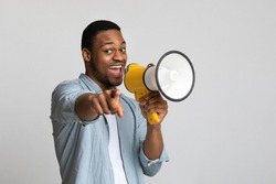Positive african american guy shouting in megaphone and pointing at camera over grey background, copy space. Happy black man screaming with loudspeaker, cheering up, making advertisement