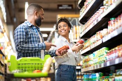 Happy African Family Couple Buying Food In Supermarket, Choosing Products Walking With Cart Along Aisles And Full Shelves Purchasing Groceries Together. Black Spouses Purchasing Essentials Together