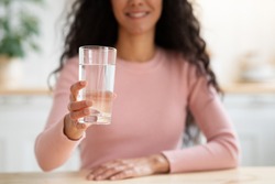 Healthy Drink. Unrecognizable Smiling Lady Offering Glass With Mineral Water At Camera While Sitting At Table In Kitchen, Brunette Woman Drinking Aqua At Home, Closeup Shot With Selective Focus