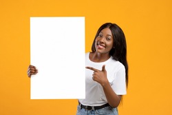 Cheerful african american young lady pointing at empty advertising placard in her hand, yellow studio background. Smiling black woman holding blank board for advertisement or text