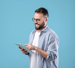 Positive young man in glasses using digital tablet for online work or studies on blue studio background. Happy teacher giving webinar. Blogger writing new post for social media